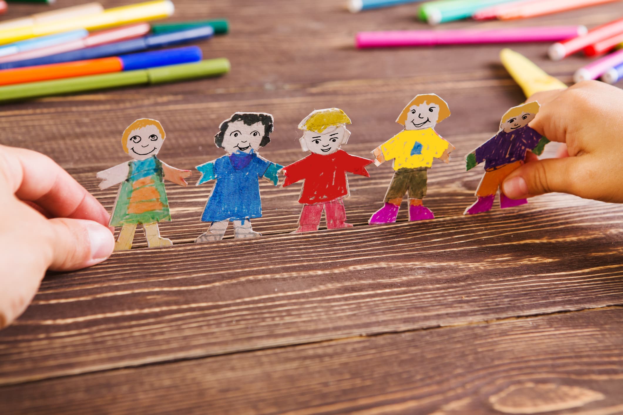 A chain of hand-drawn paper dolls held up on a wooden desk beside colored markers